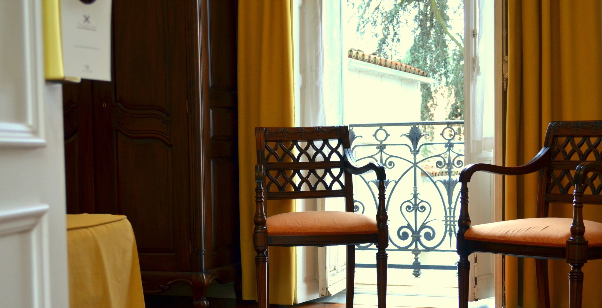 Une pièce avec deux chaises en bois dotées d’assises rembourrées, une porte en bois et une fenêtre ornée de rideaux jaunes.