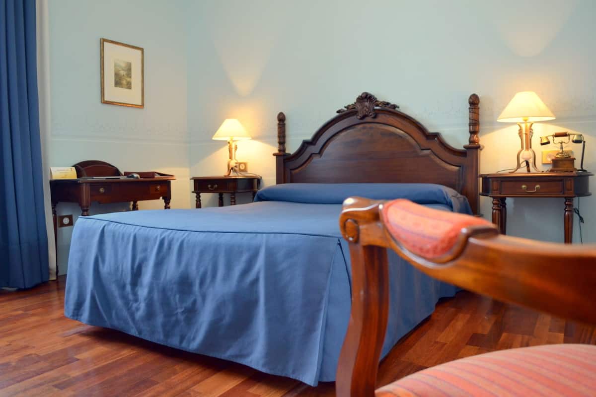 A neatly made bed with a blue bedspread, two bedside tables with lamps, a wooden chair, and a framed picture on the wall.