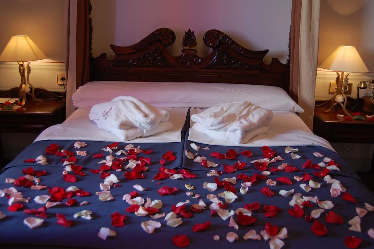 A bed with rose petals scattered across the bedspread, two white towels folded on the bed, and a wooden headboard.