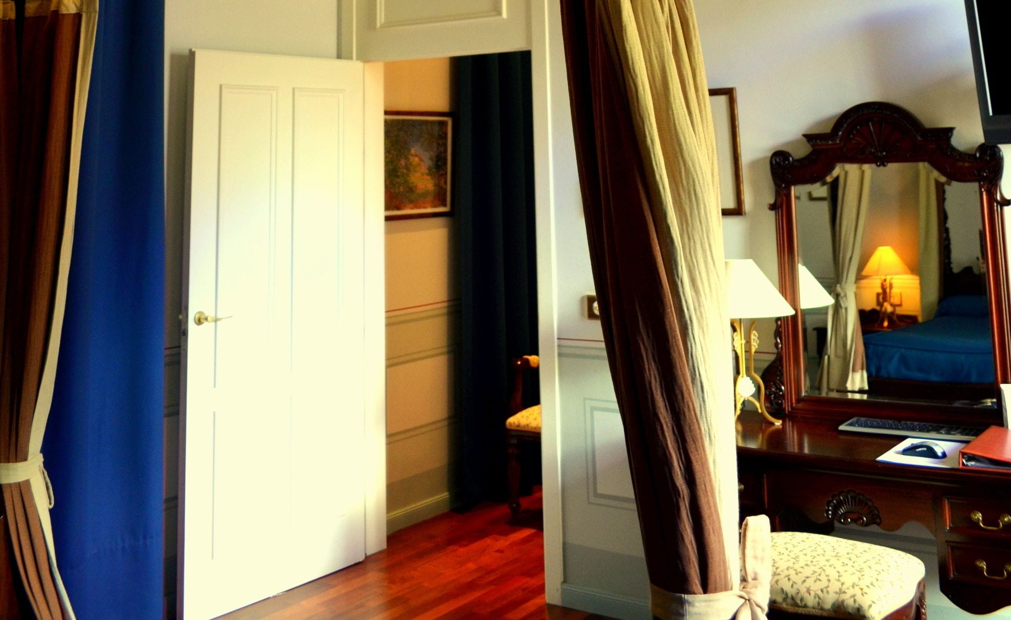A bedroom with a wooden floor, a bed with a blue bedspread, a desk with a lamp, a chair, and a large mirror.