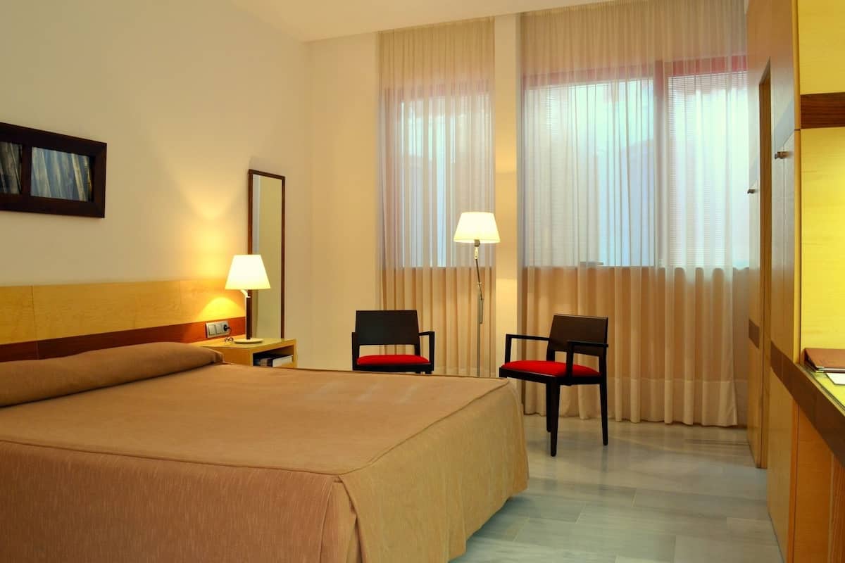 A hotel room with a large bed, two chairs, a desk, and a window with curtains.