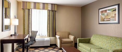 Quarto de hotel com escrivaninha, duas cadeiras, um sofá, uma mesinha e uma janela com cortinas.