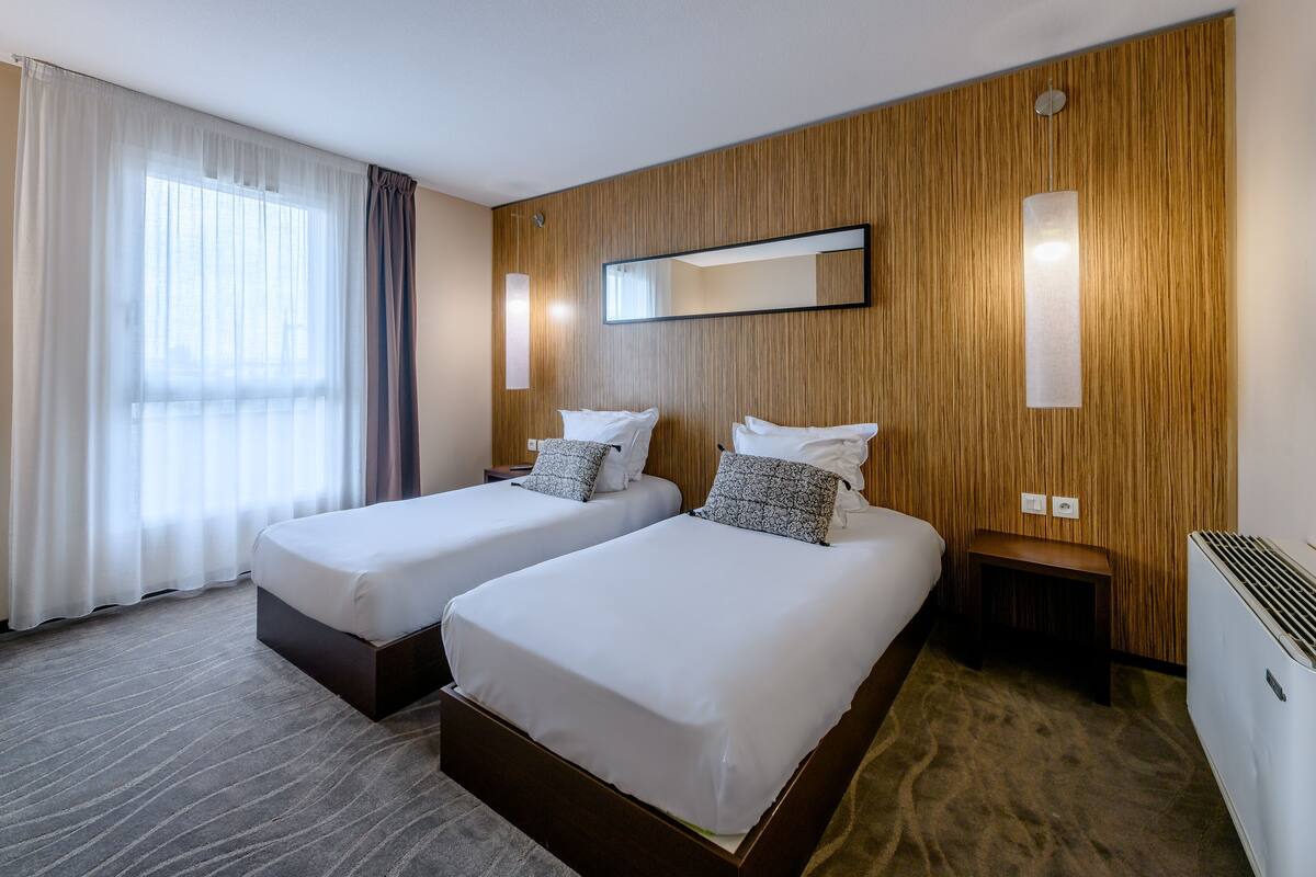Une chambre d’hôtel avec deux lits, une grande fenêtre avec des rideaux, un mur décoratif en bois, un miroir et une table de chevet.