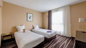 A hotel room with two beds, a striped carpet, a nightstand with a lamp, and a window with curtains.