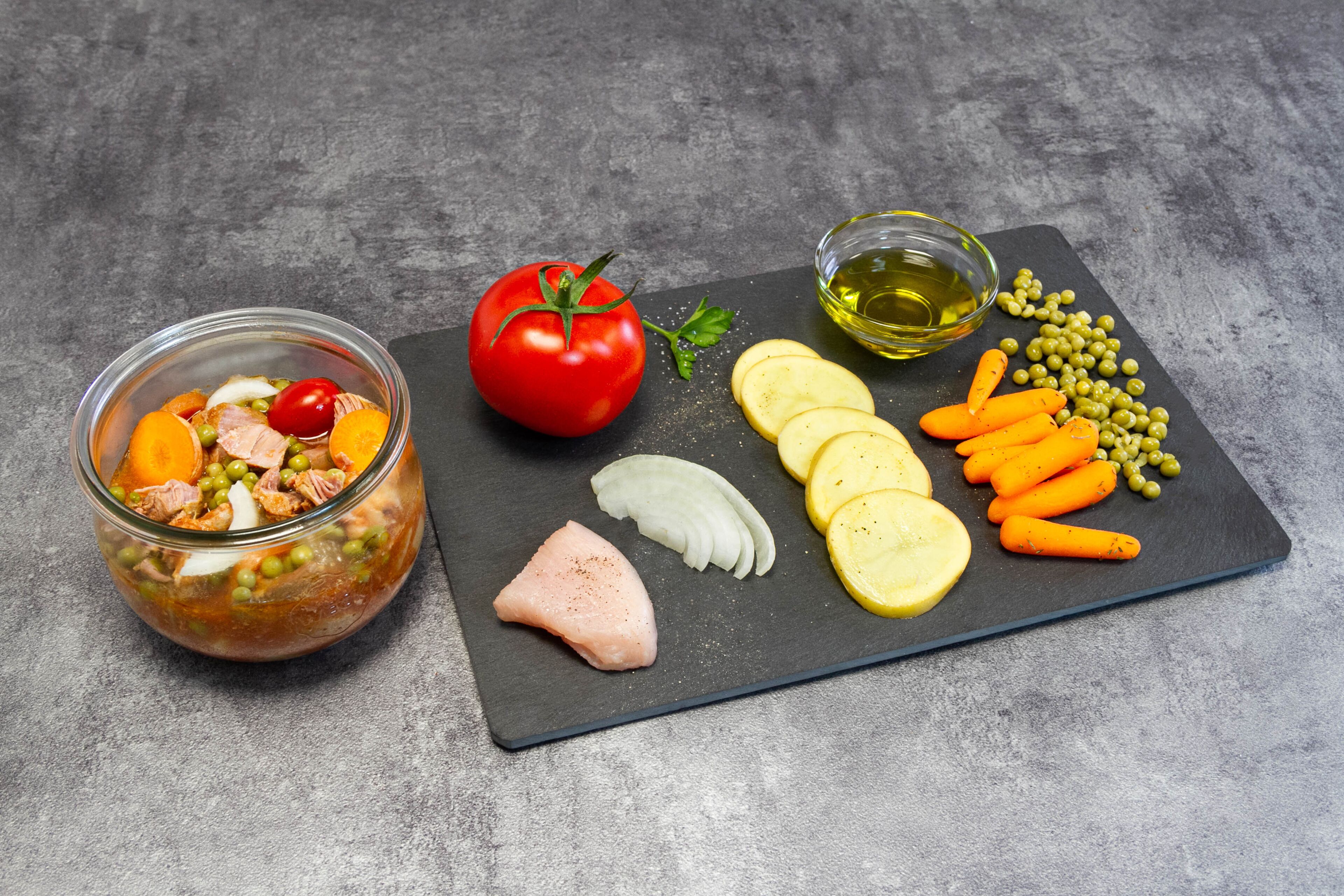 Un saladier en verre contenant un mélange d’ingrédients, une tomate entière, des légumes coupés en tranches et un petit bol d’huile, le tout posé sur une planche d’ardoise.