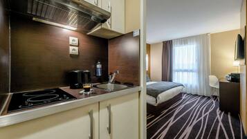 A compact hotel room with a kitchenette, a bed, a desk, and a window with curtains.
