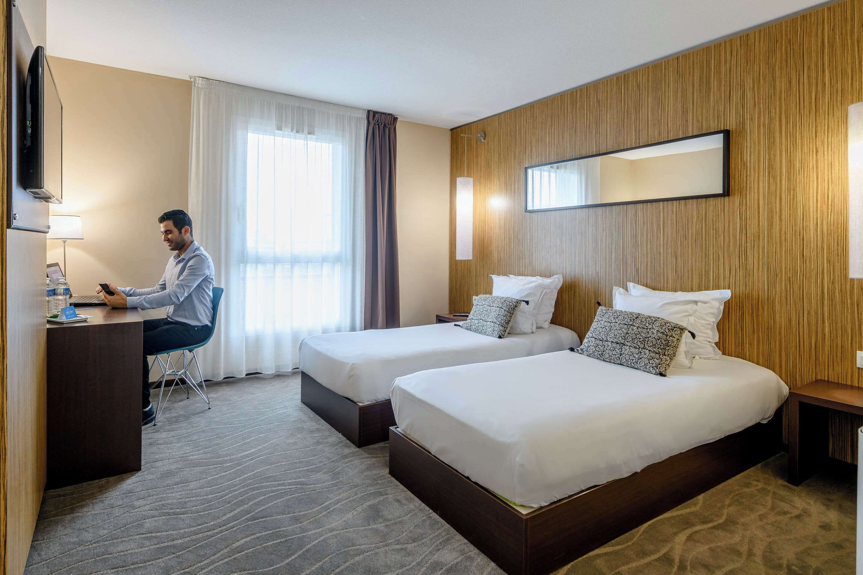 Une chambre d’hôtel avec deux lits, un bureau, une télévision et une grande fenêtre avec des rideaux.