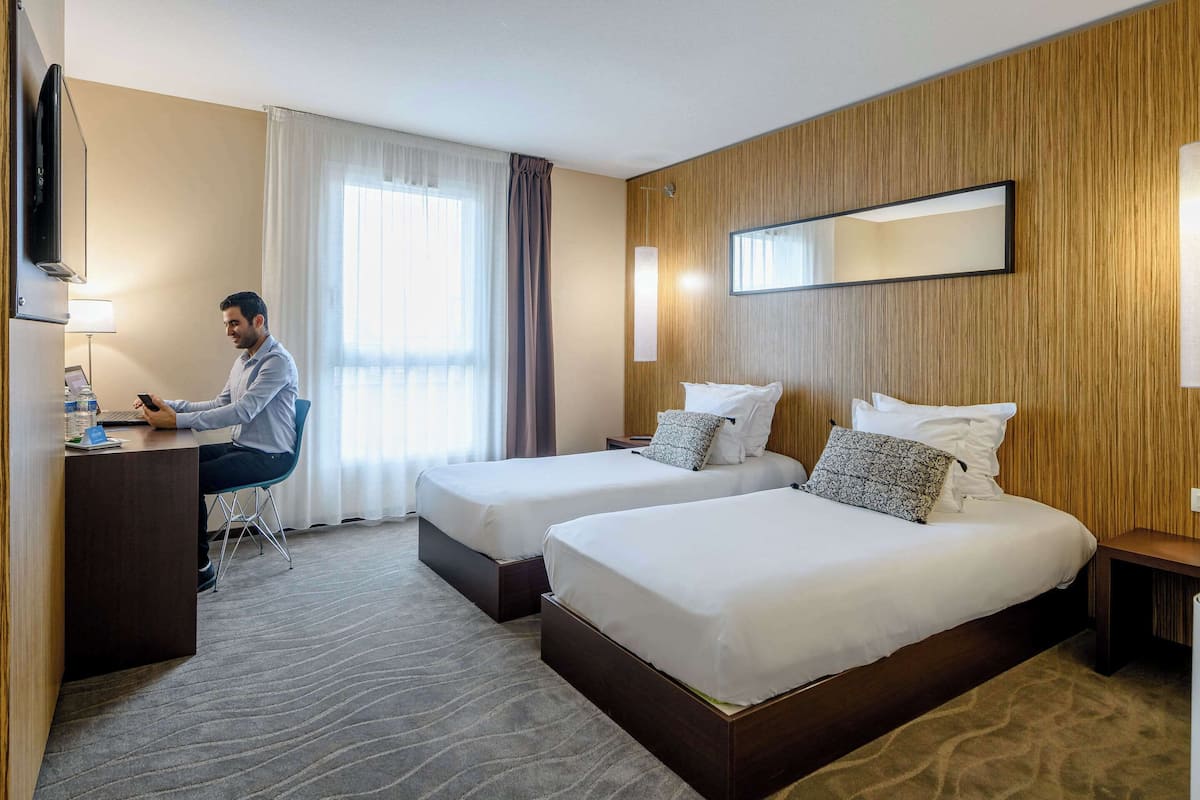 Une chambre d’hôtel avec deux lits, un bureau, une télévision et une grande fenêtre avec des rideaux.