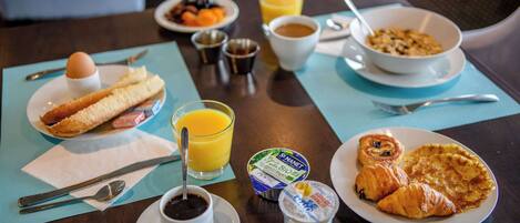 Une table de petit-déjeuner avec des croissants, un bol de céréales, un verre de jus d’orange, une tasse de café et une assiette de fruits.