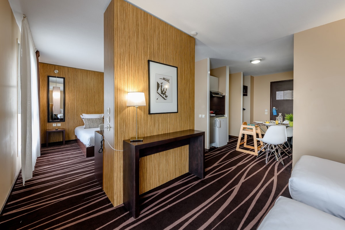 Ein Hotelzimmer mit einer Wandpaneinung aus Holz, einem Bett, einem Schreibtisch, einem Stuhl, einem kleinen Tisch, einer Lampe, einem gerahmten Bild, einem Spiegel, einer Küchenzeile und einem Teppichboden.