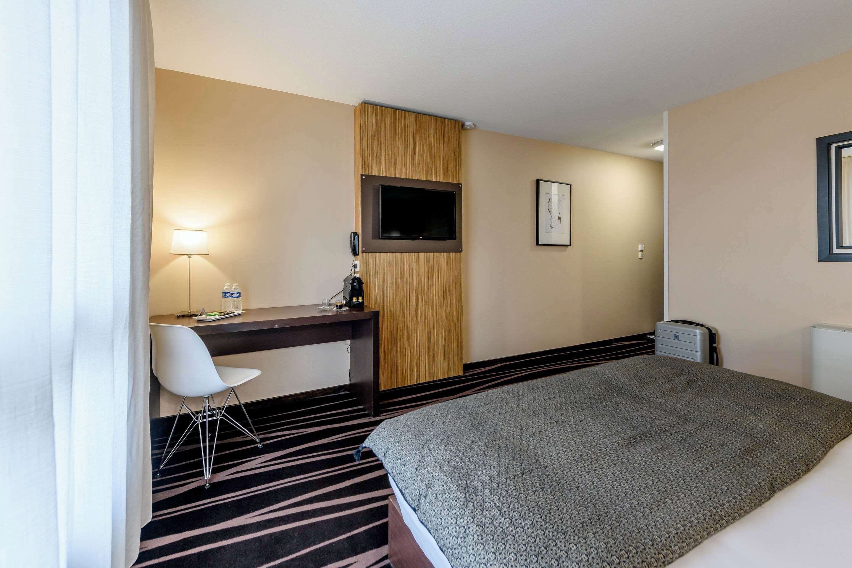 A hotel room with a bed, a desk with a chair, a flat-screen TV, a lamp, and a small safe.