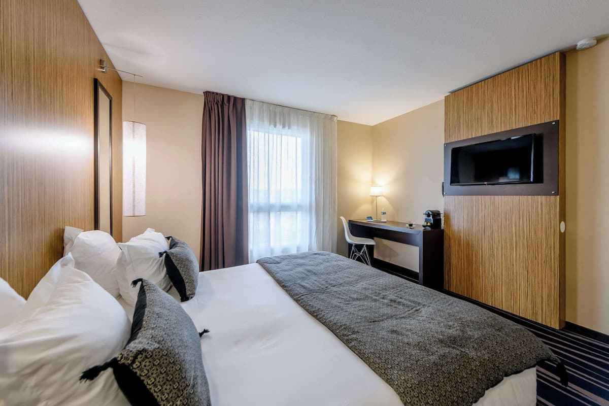 Une chambre d’hôtel avec un grand lit, un bureau avec une lampe, un téléviseur fixé au mur et une fenêtre avec des rideaux.