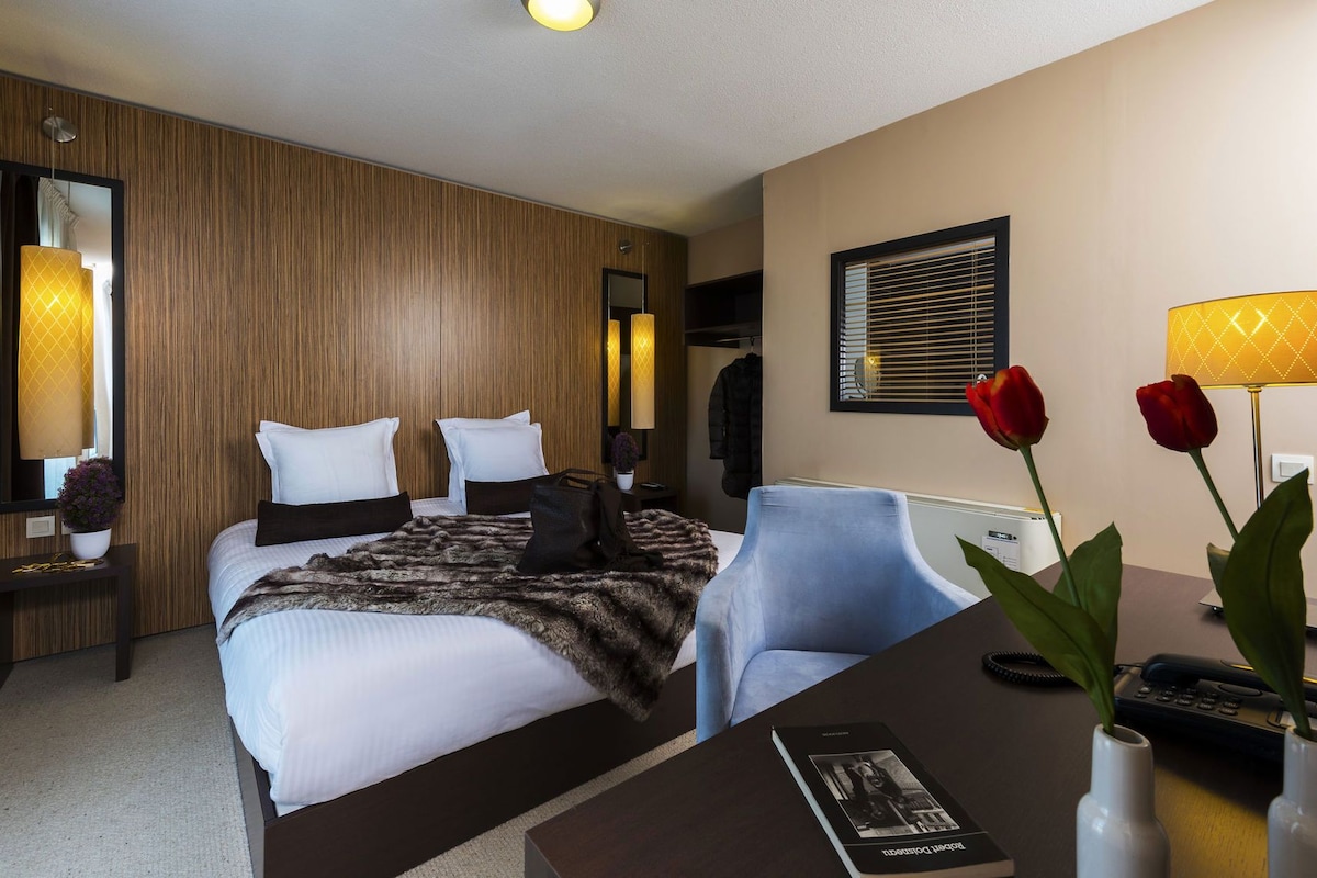 Ein Hotelzimmer mit einem Bett, einem Schreibtisch, einem Sessel, einem Spiegel, einer Lampe und einer Vase mit Blumen.