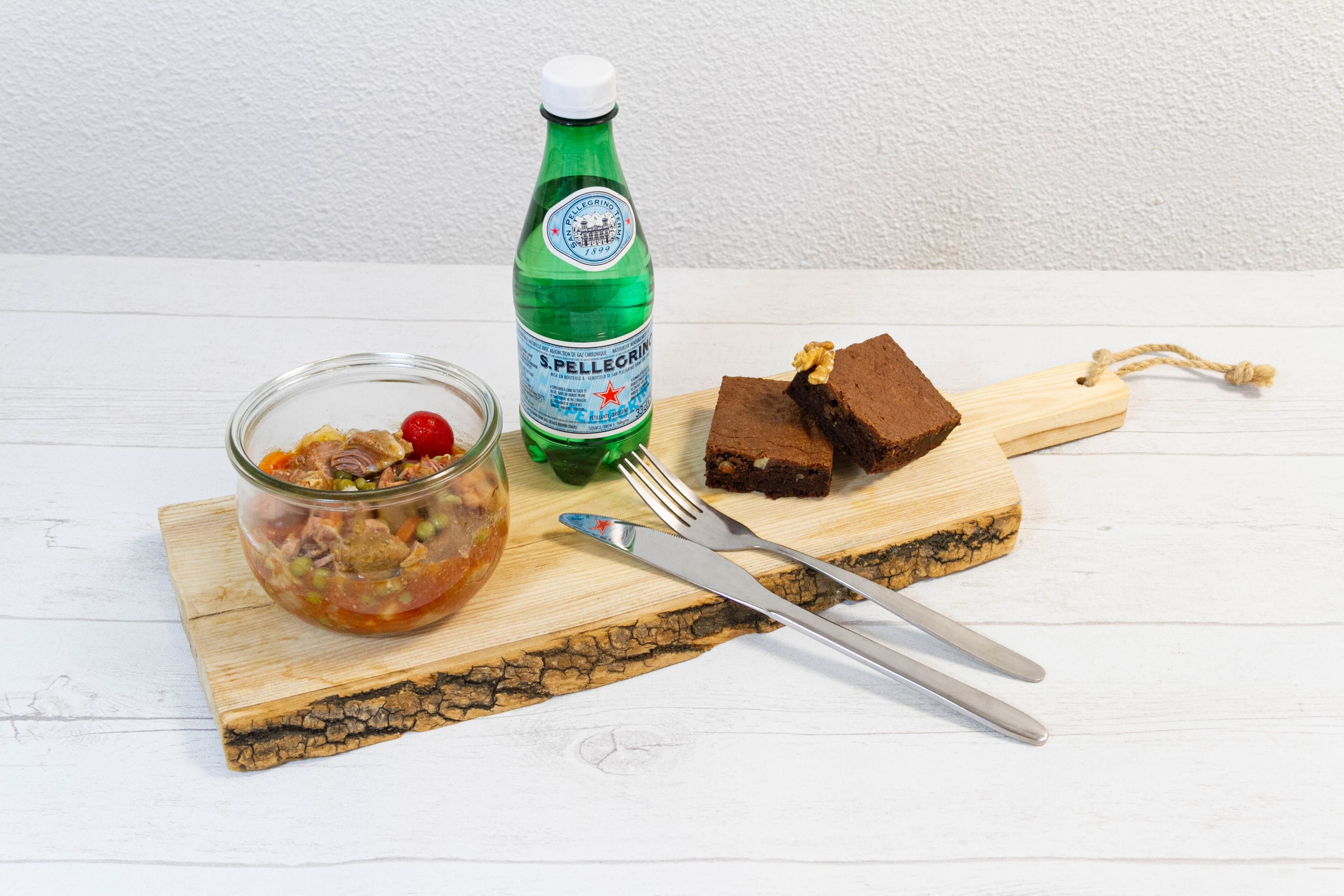 A wooden cutting board with a glass jar of pickled vegetables, a bottle of S.Pellegrino water, and two pieces of chocolate cake.