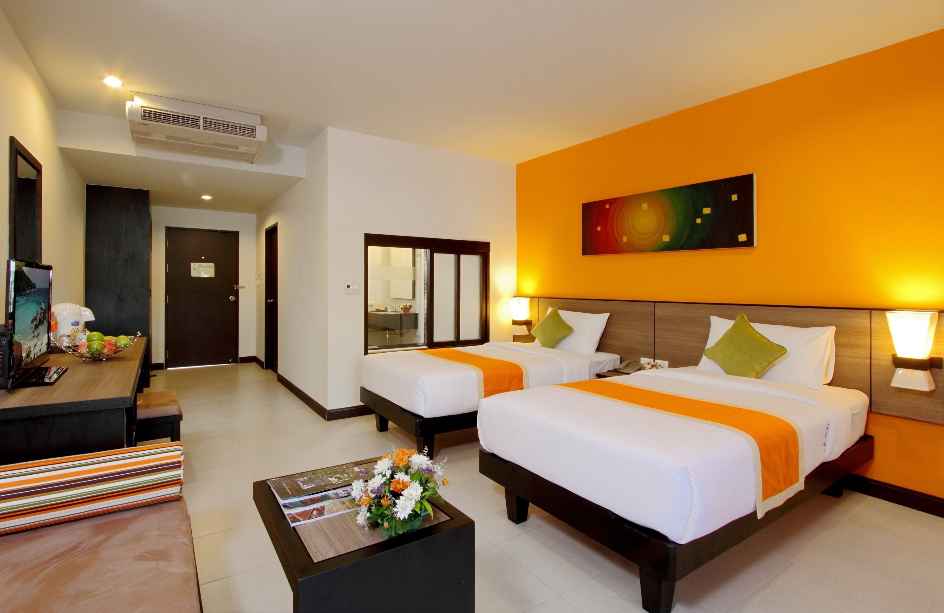 A hotel room with two beds, a desk with a computer, a TV, and a bathroom visible through a glass door.
