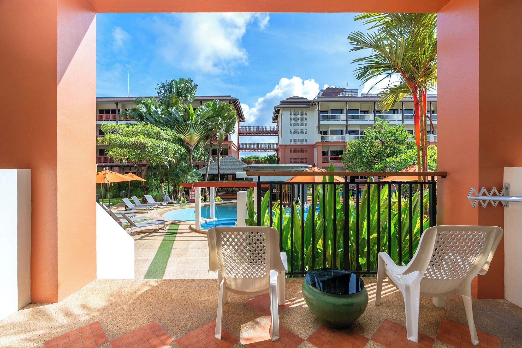 Un balcón con dos sillas blancas, una piscina y vista a un edificio de hotel con balcones y palmeras.