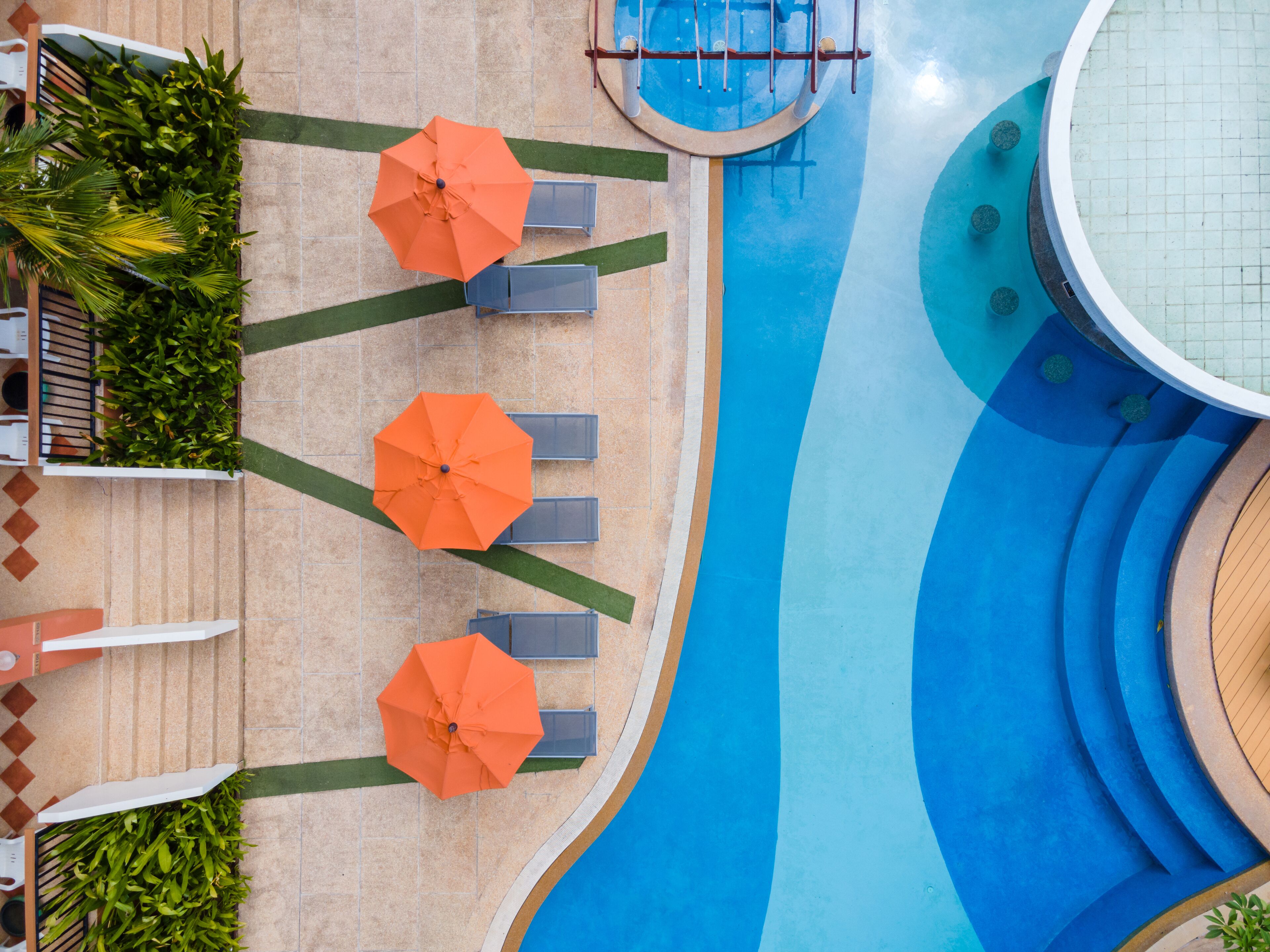 An aerial view of a swimming pool area with three orange umbrellas, lounge chairs, and a tiled walkway.