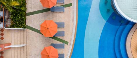 An aerial view of a swimming pool area with three orange umbrellas, lounge chairs, and a tiled walkway.