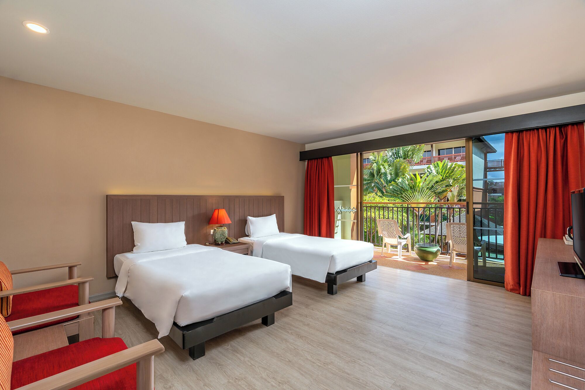 A hotel room with two beds, a TV, a desk, and a balcony with a view of a pool and palm trees.