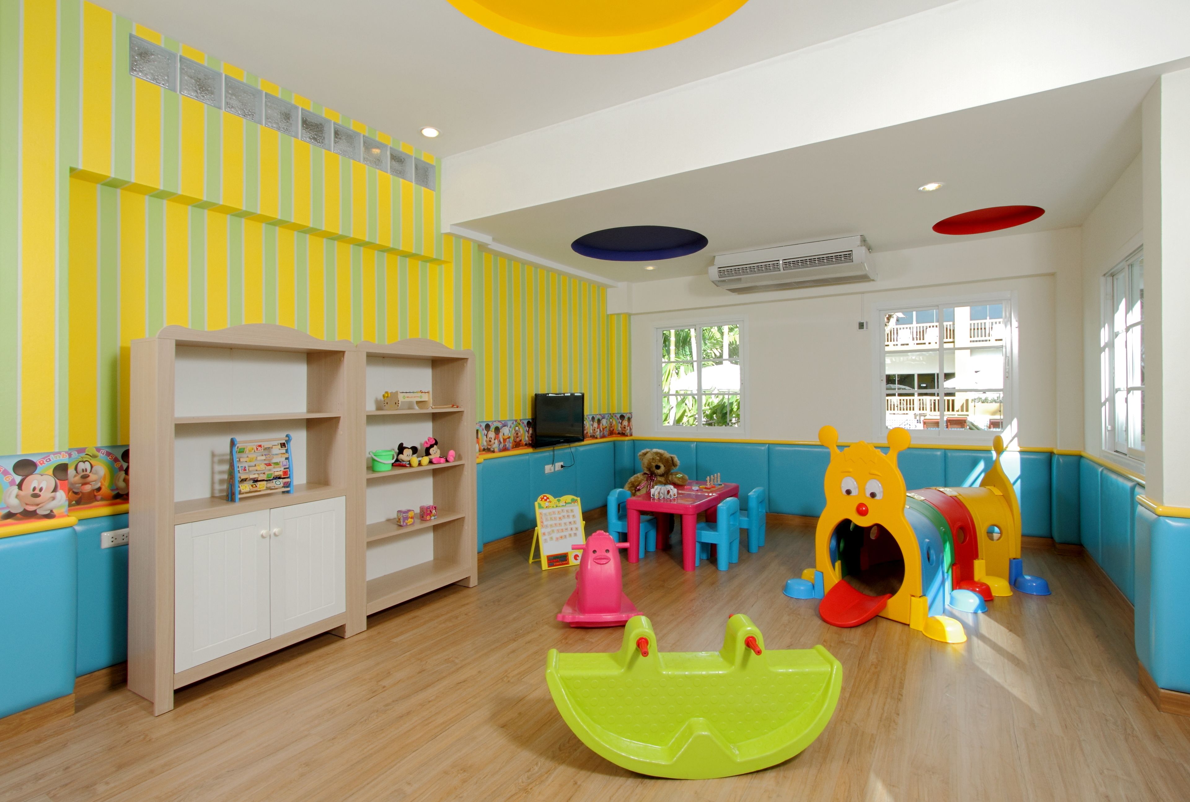 A brightly colored children's playroom with a wooden floor, a bookshelf, a teddy bear, and a play area with a slide and a tunnel.