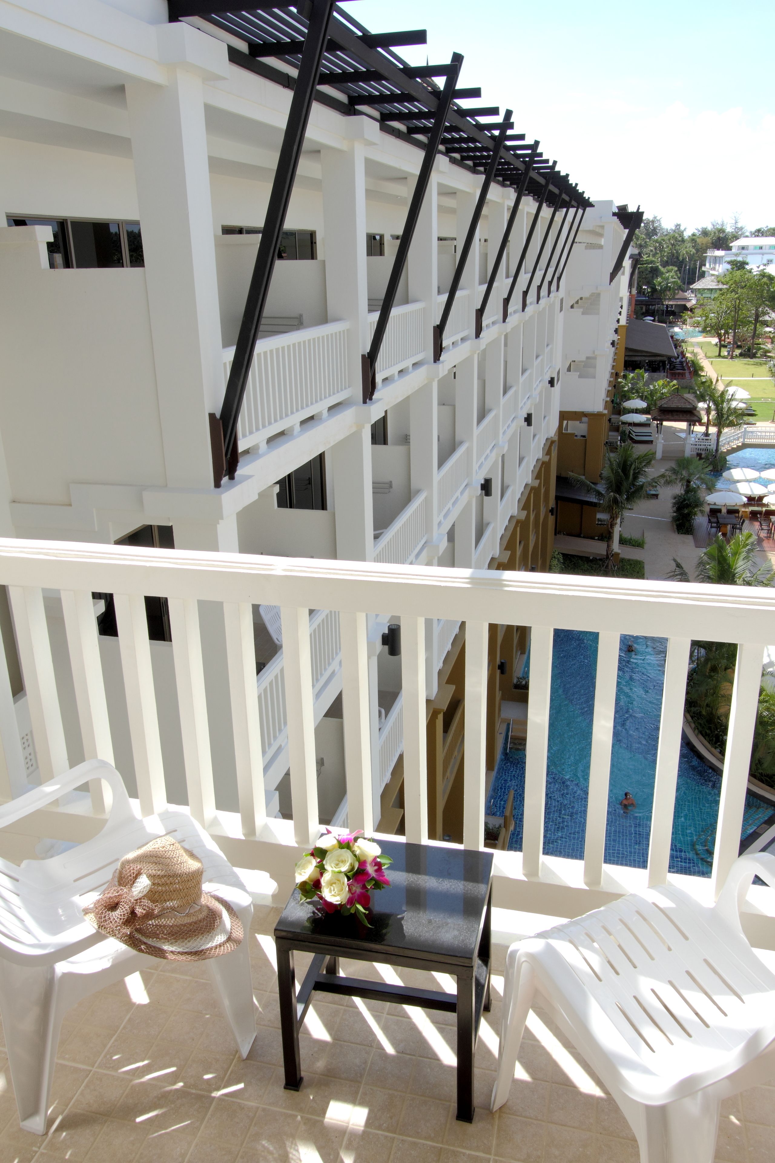 Un balcón con sillas de plástico blanco, una pequeña mesa con un jarrón de flores y vistas a una piscina y edificios de un hotel.
