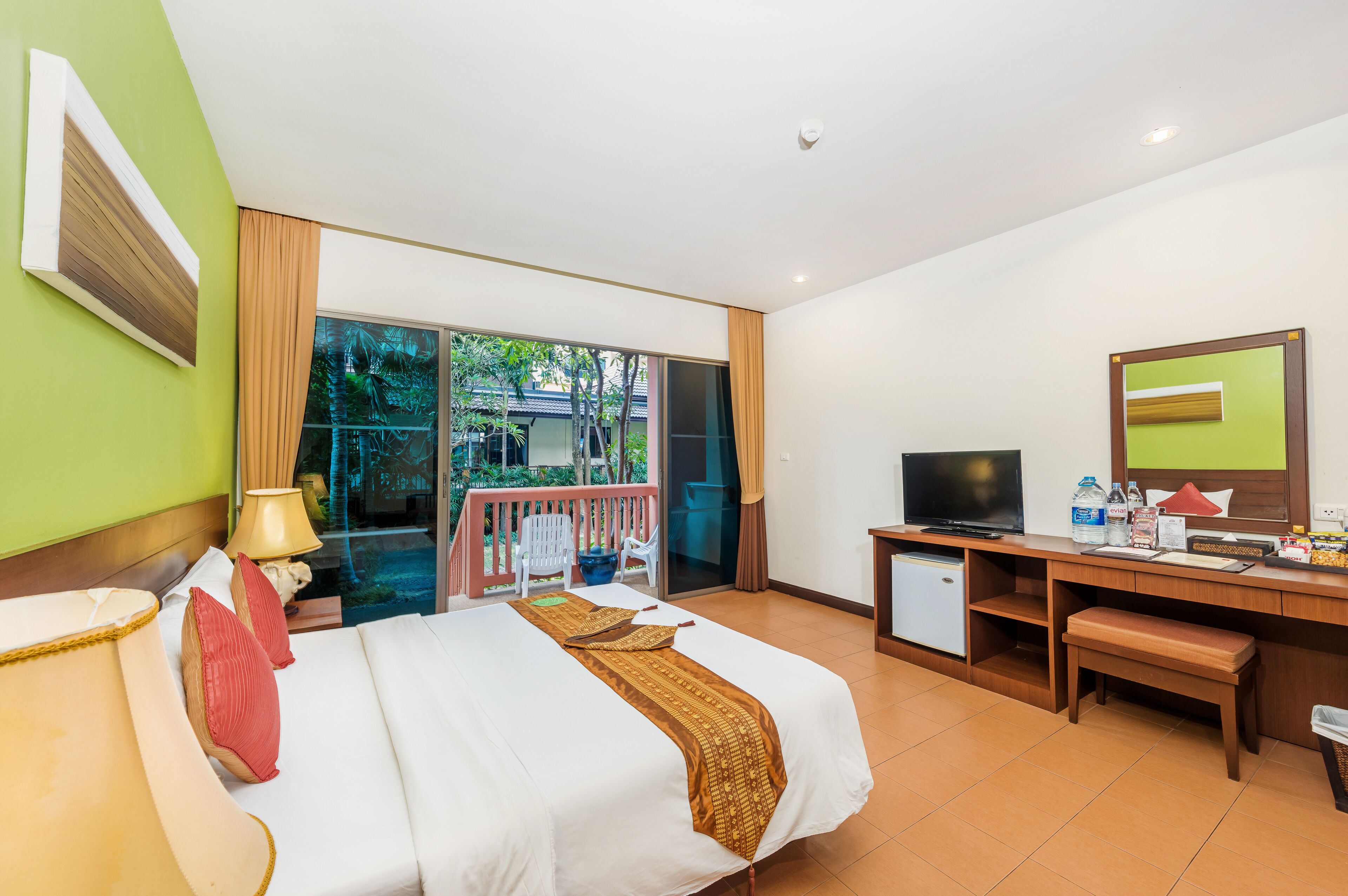 A hotel room with a bed, desk, TV, and a balcony with a view of greenery.