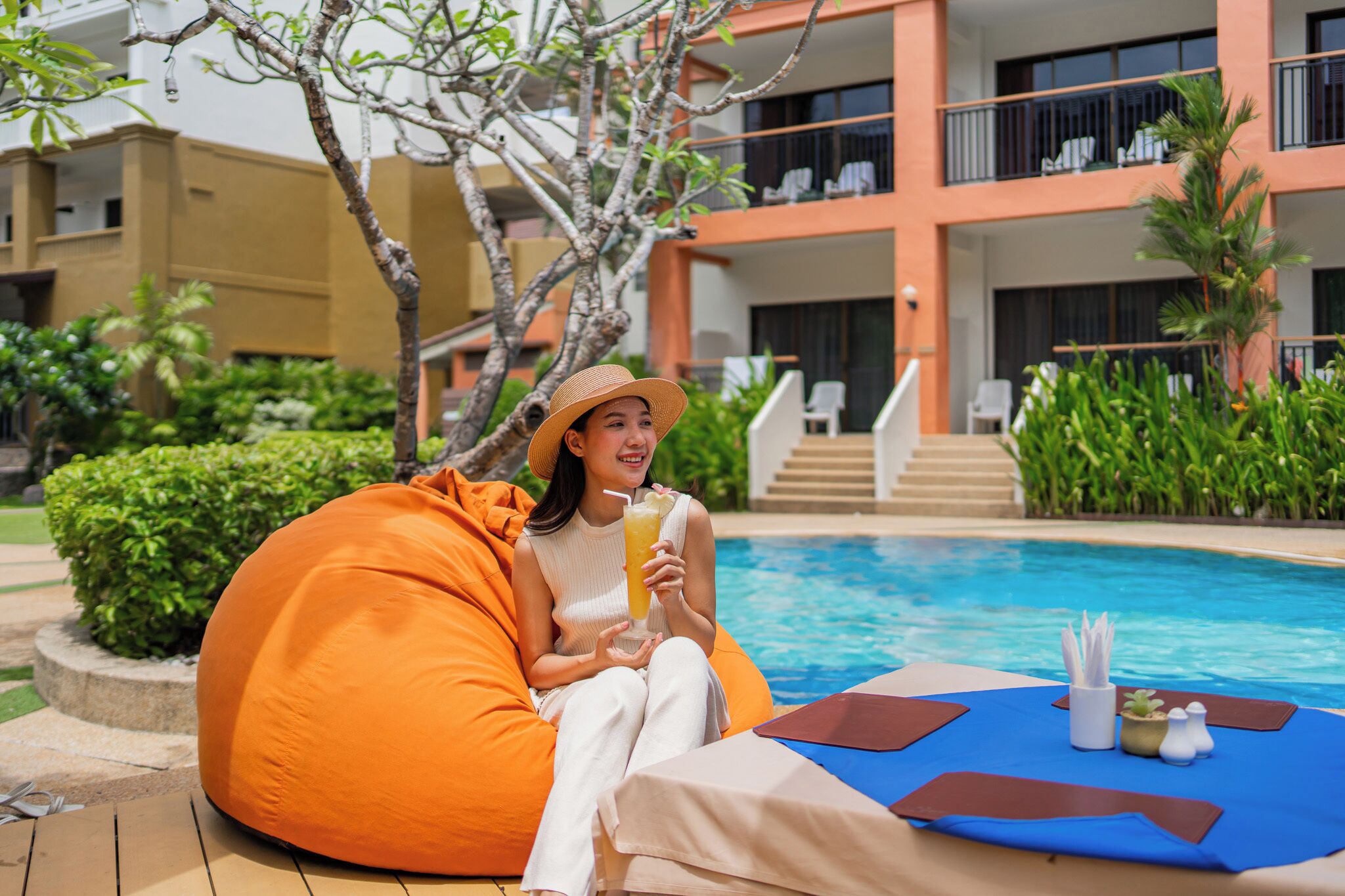 A person sitting on an orange bean bag chair by a poolside with a drink, surrounded by a pool, greenery, and a building with balconies.