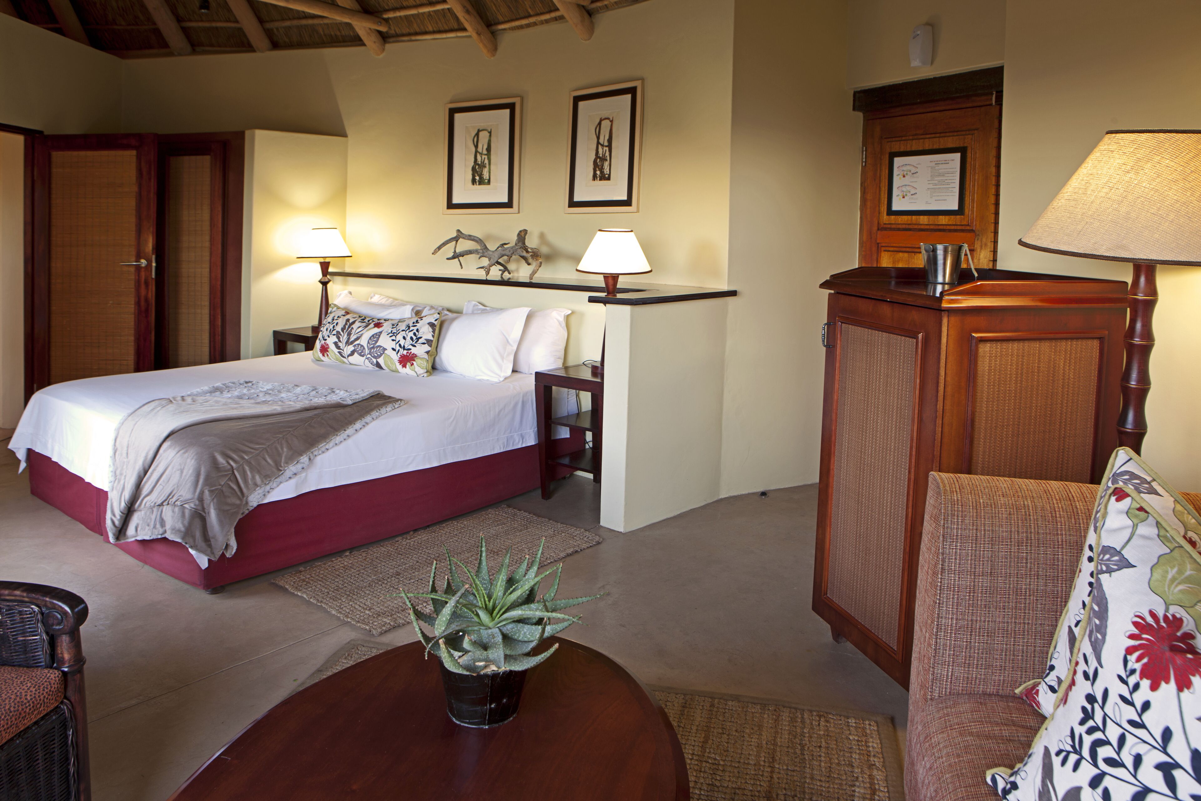 A bedroom with a bed, bedside lamps, a nightstand, a chair, a table with a plant, and a door.