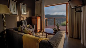 A cozy living room with a sofa, a coffee table, and a TV mounted on the wall. There are framed pictures on the wall and a view of the outdoors through a large window. - Kuzuko Lodge (Blue Crane Route)