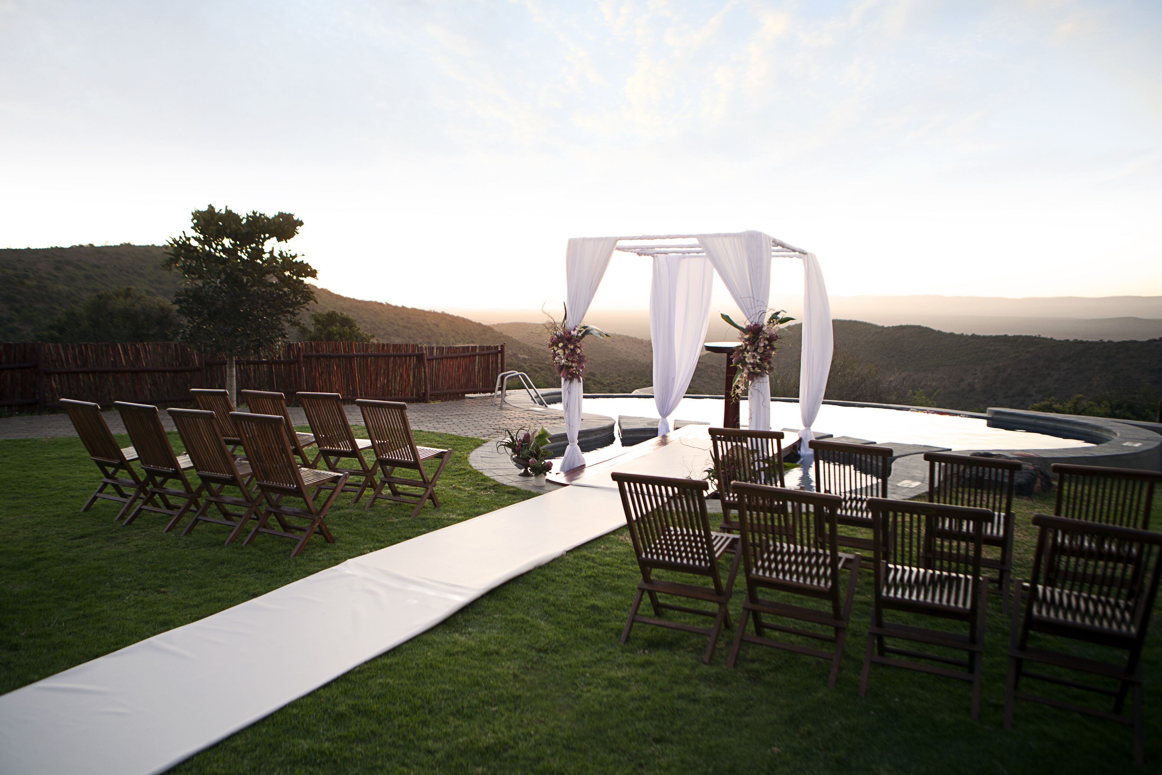 Montaje de boda al aire libre con un arco blanco y sillas dispuestas sobre una zona cubierta de césped, con vistas a colinas.