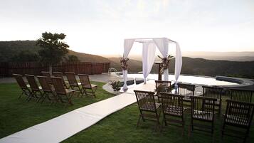 An outdoor wedding setup with a white arch and chairs arranged on a grassy area with a view of hills.