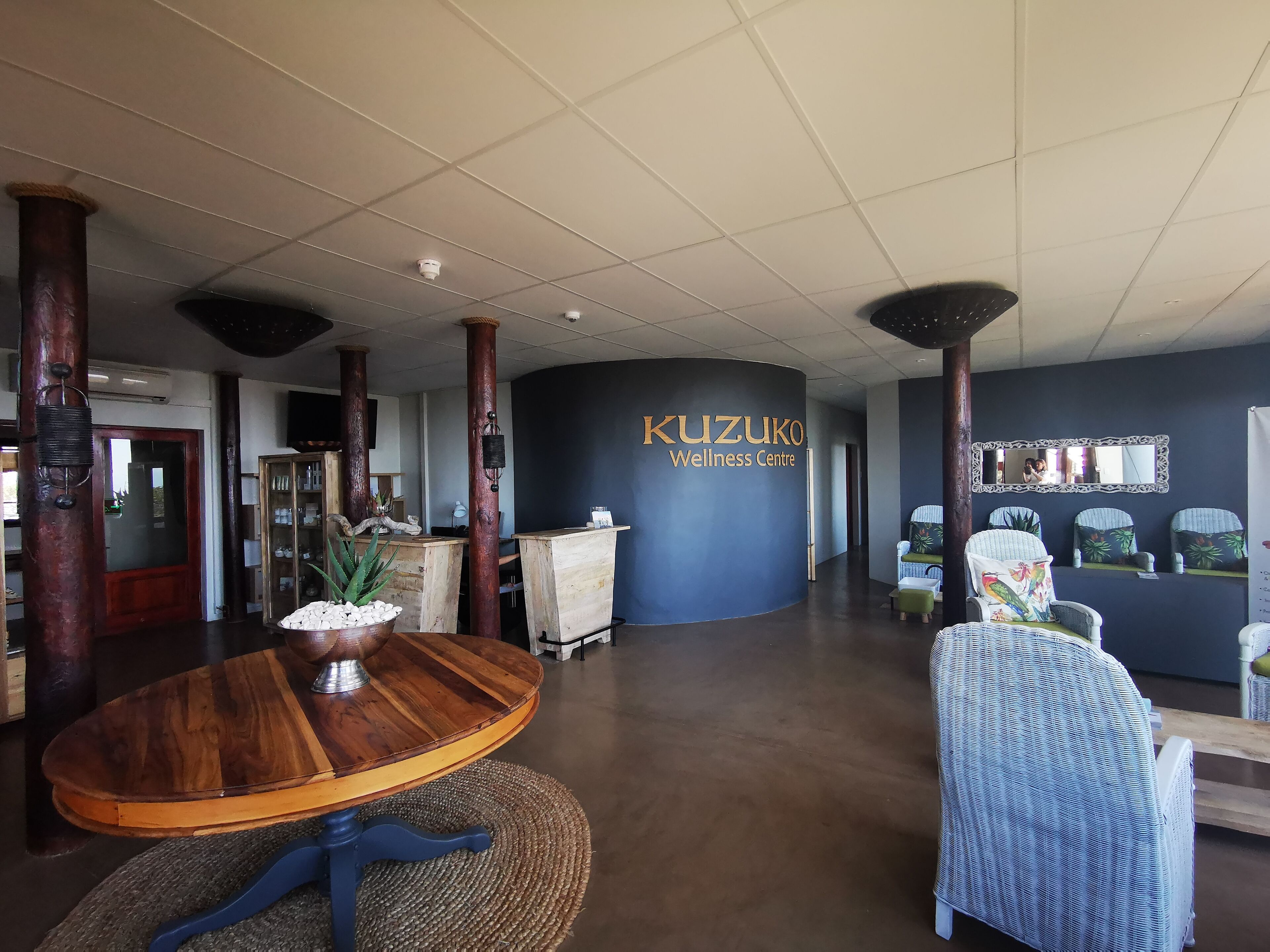A reception area with a wooden reception desk, a round wooden table with a woven mat, and a blue wall with the text "KUZUKO Wellness Centre".