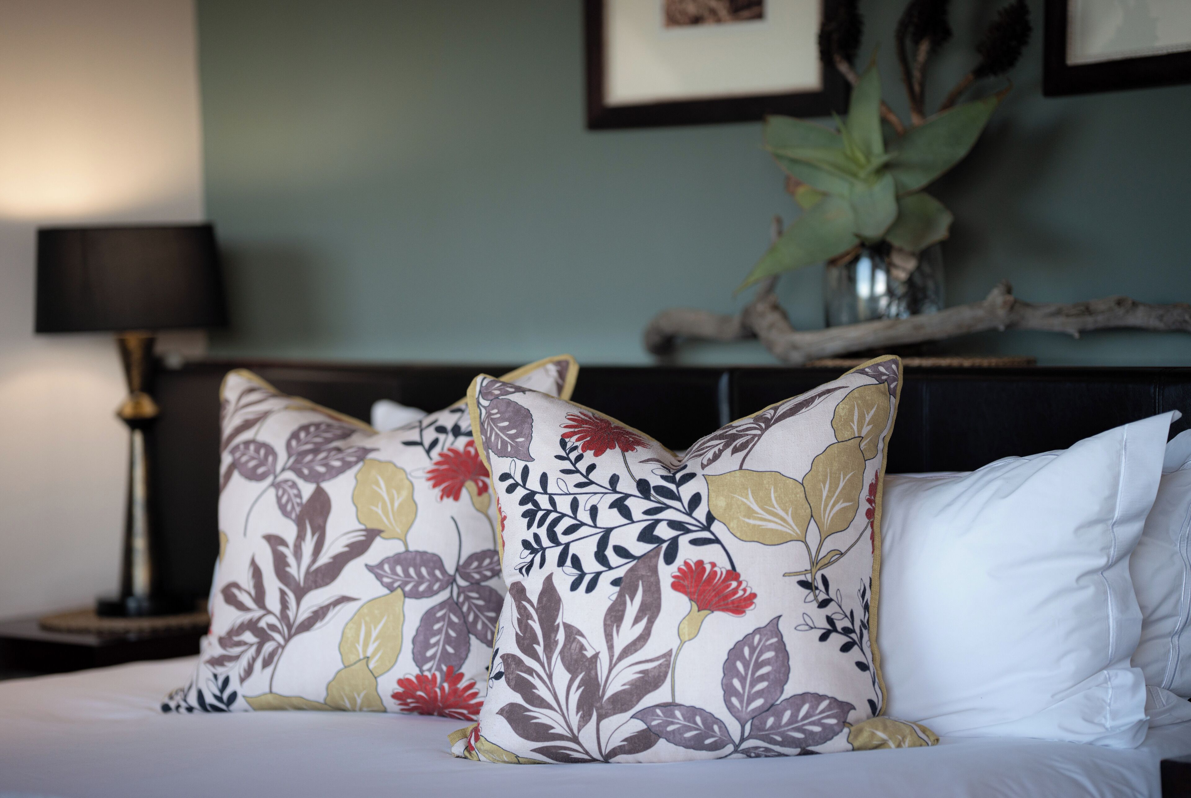 A bed with floral-patterned pillows and a white pillow, a lamp, and a plant on a nightstand.