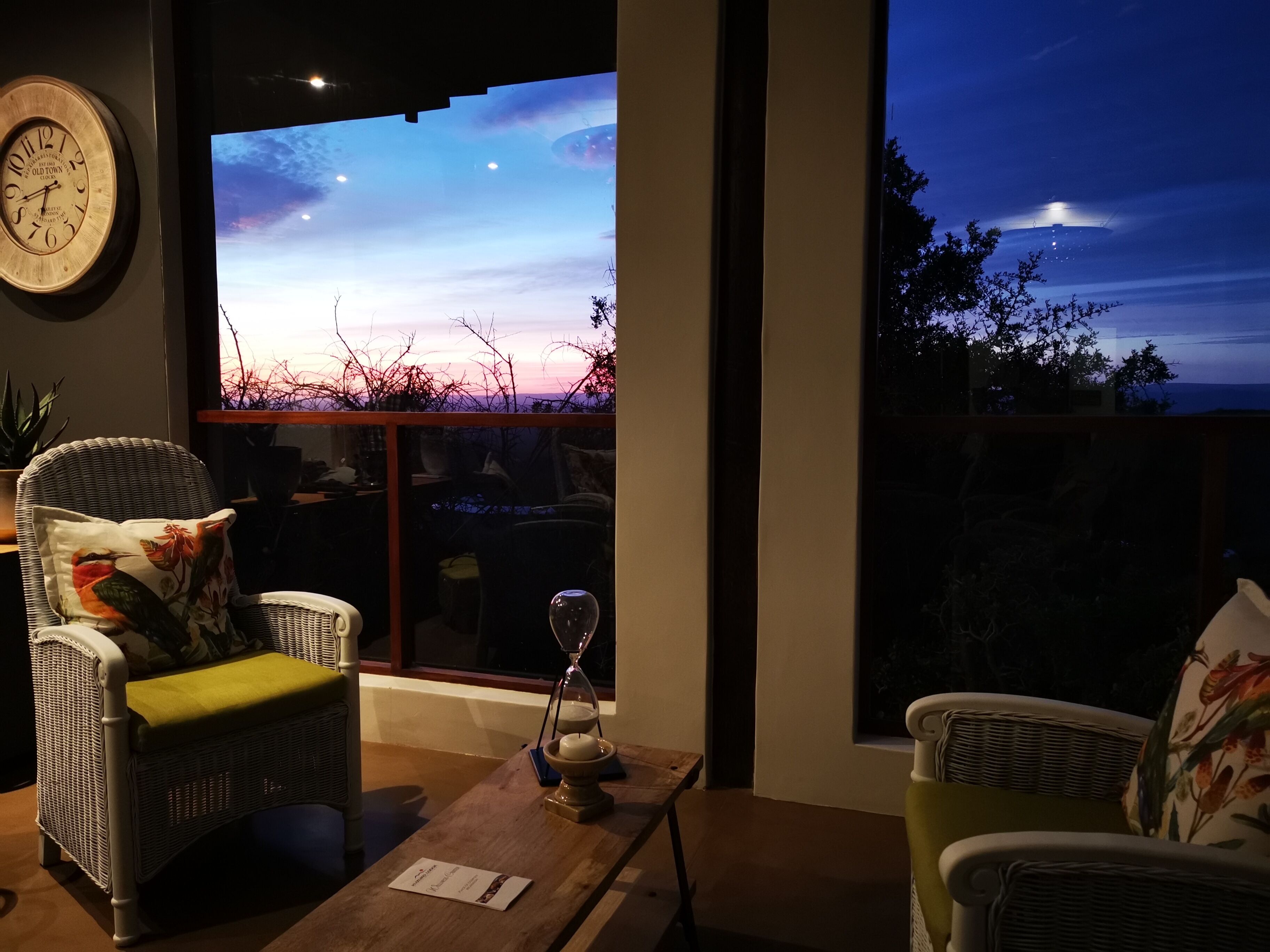 A room with wicker chairs, a wooden table, and a large window displaying a sunset view.