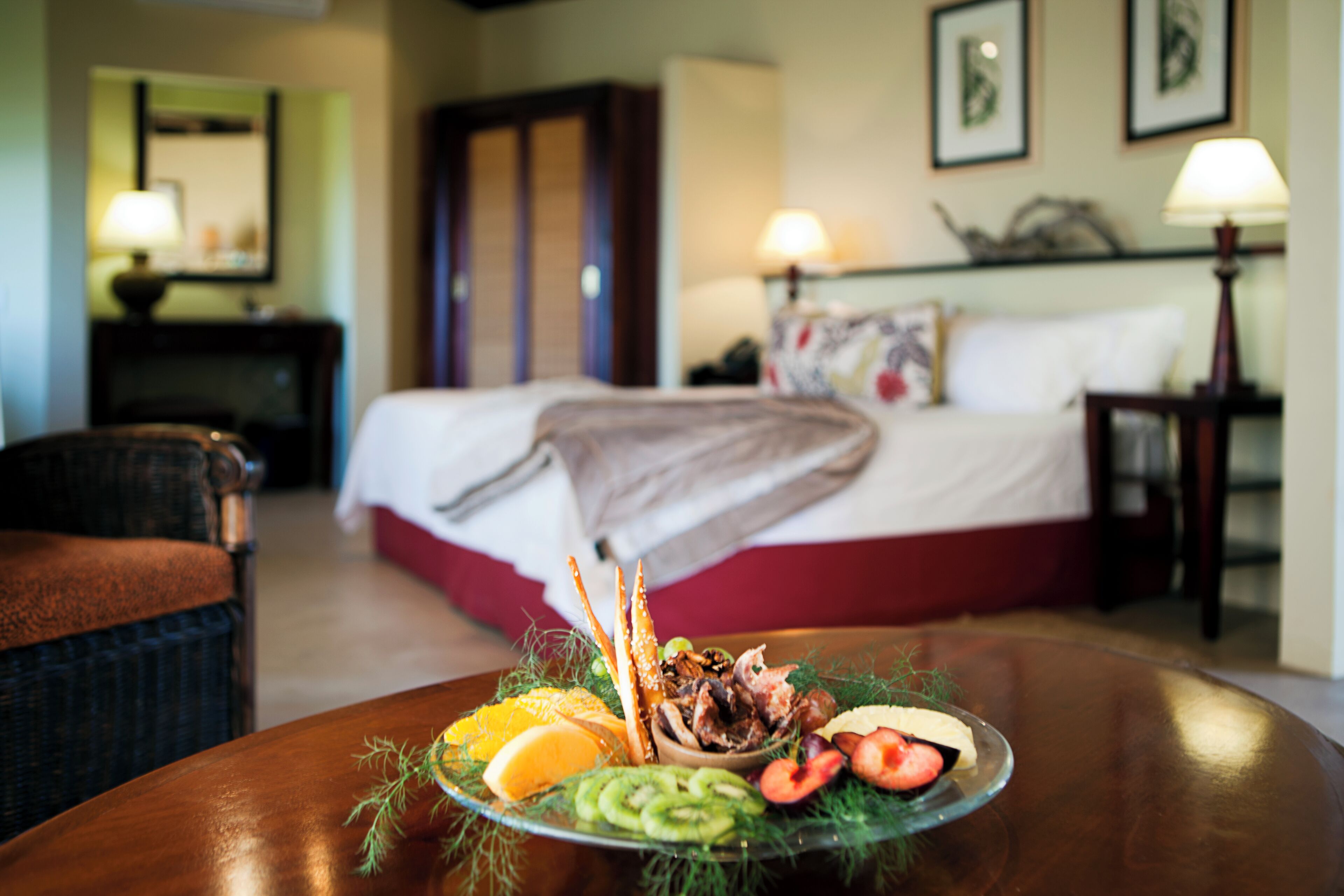 A hotel room with a bed, a dining table with a fruit and vegetable platter, a chair, and a lamp.