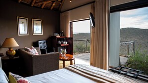 A hotel room with a bed, a desk, a chair, a bookshelf, and a large window offering a view of the outdoors. - Kuzuko Lodge (Blue Crane Route)