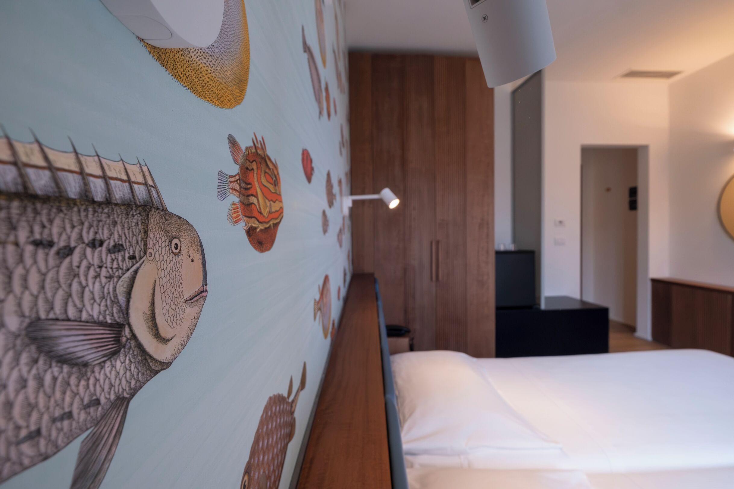 Ein Schlafzimmer mit einem Bett, einer Wand mit Fischillustrationen, einem hölzernen Kopfteil, einem Nachttisch und einer Lampe.