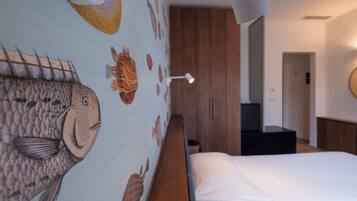 A bedroom with a bed, a wall with fish illustrations, a wooden headboard, a bedside table, and a lamp.