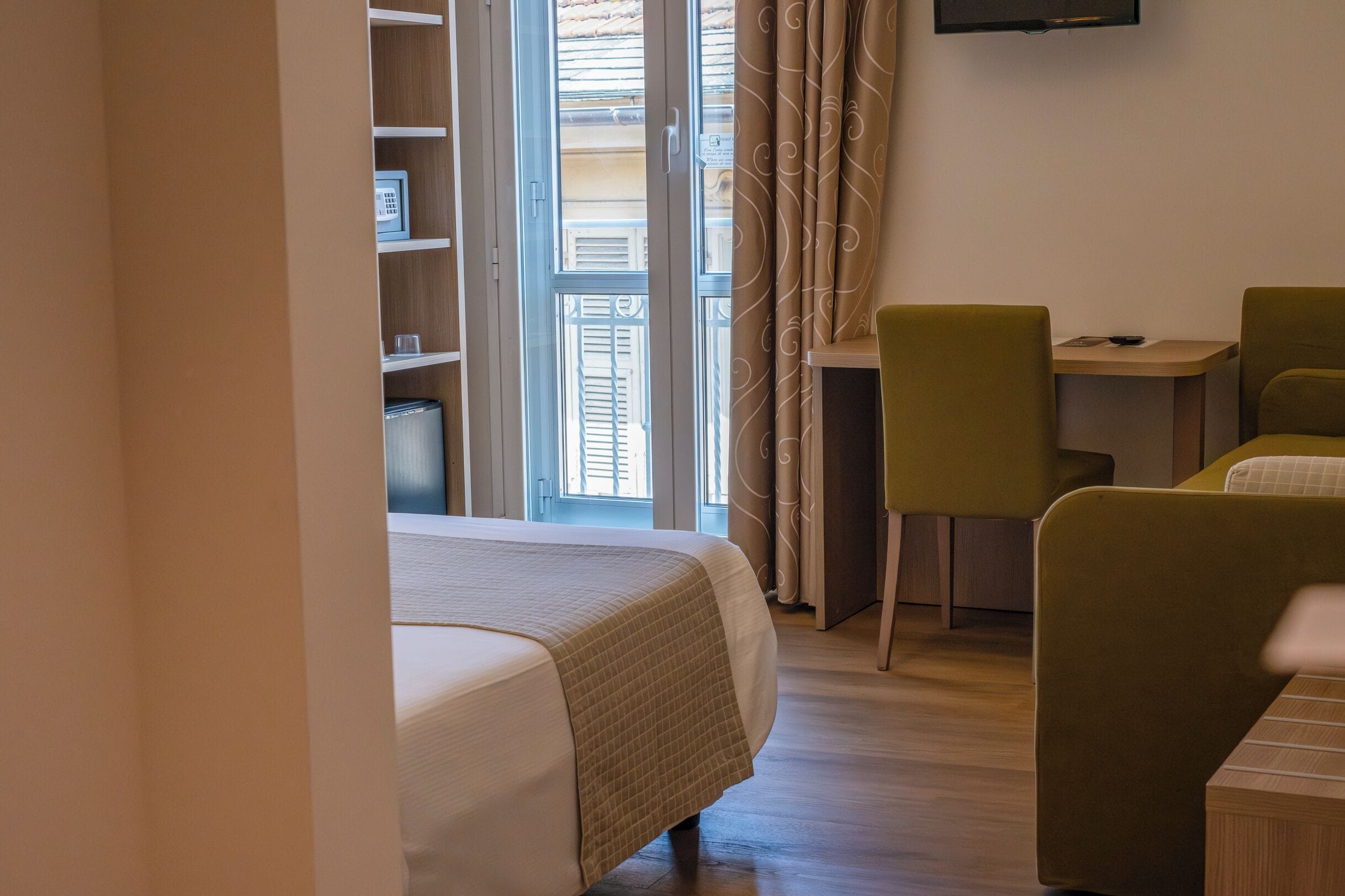 A hotel room with a bed, a desk, two chairs, a TV mounted on the wall, and a window with curtains.