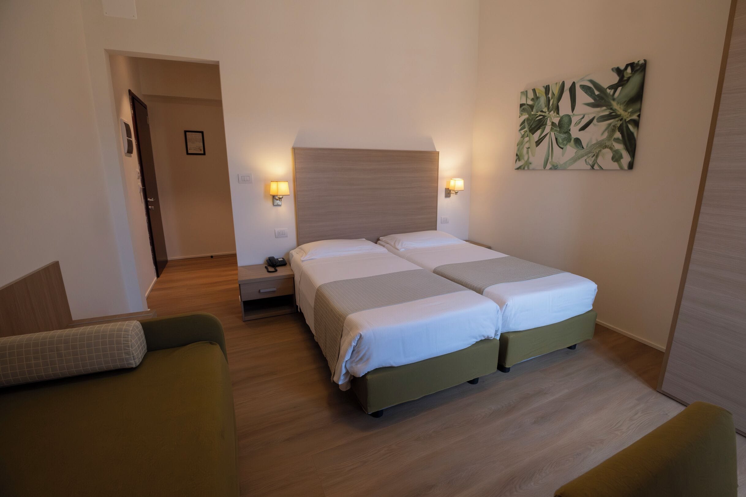 Habitación de hotel con dos camas, un sofá, una mesita de noche y una lámpara de pared.