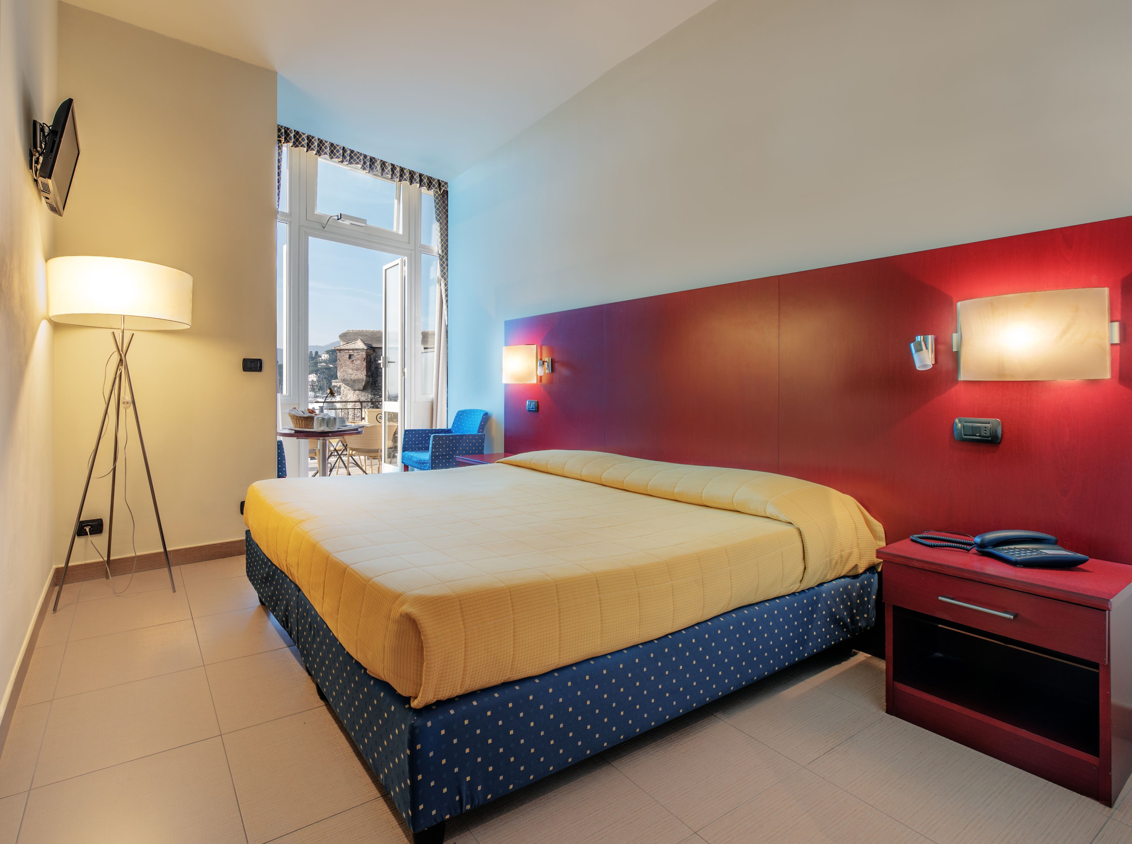 Camera d'albergo con un letto, comodino, lampada e vista sulla città attraverso una finestra.