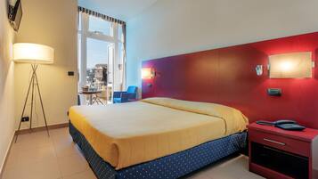 Camera d'albergo con un letto, comodino, lampada e vista sulla città attraverso una finestra.