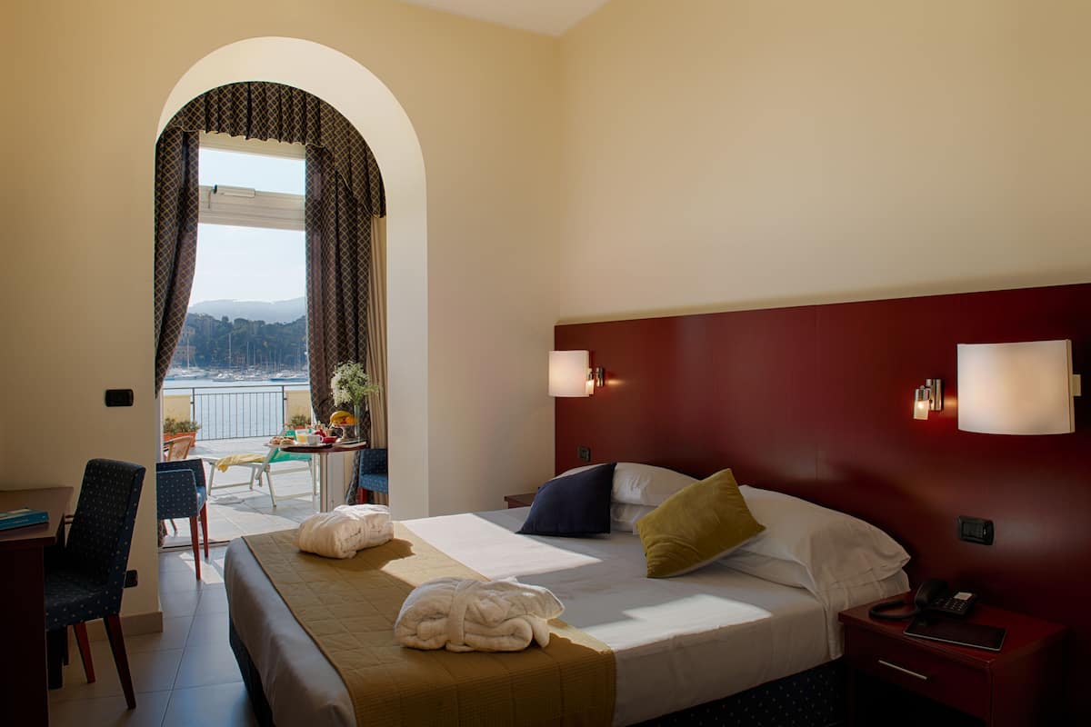 Une chambre d’hôtel avec un lit, un bureau, une chaise, une table de chevet et une vue sur la mer, visible à travers une fenêtre en arc de cercle.