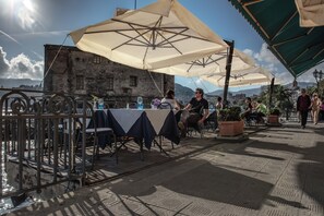 Un café en plein air avec des tables et des chaises placées sous de grands parasols, une rambarde en métal et un bâtiment en arrière-plan.