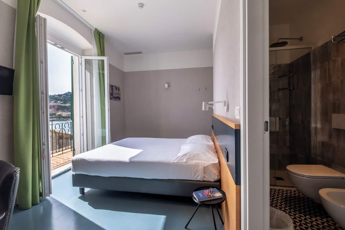 Une chambre avec un lit, une petite table de chevet, une salle de bain avec des toilettes et une douche, et un balcon avec des rideaux verts.