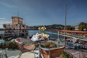 Un balcon avec des fauteuils en osier, une table où sont disposés des éléments pour le petit-déjeuner, et une vue sur un bâtiment historique et la côte.