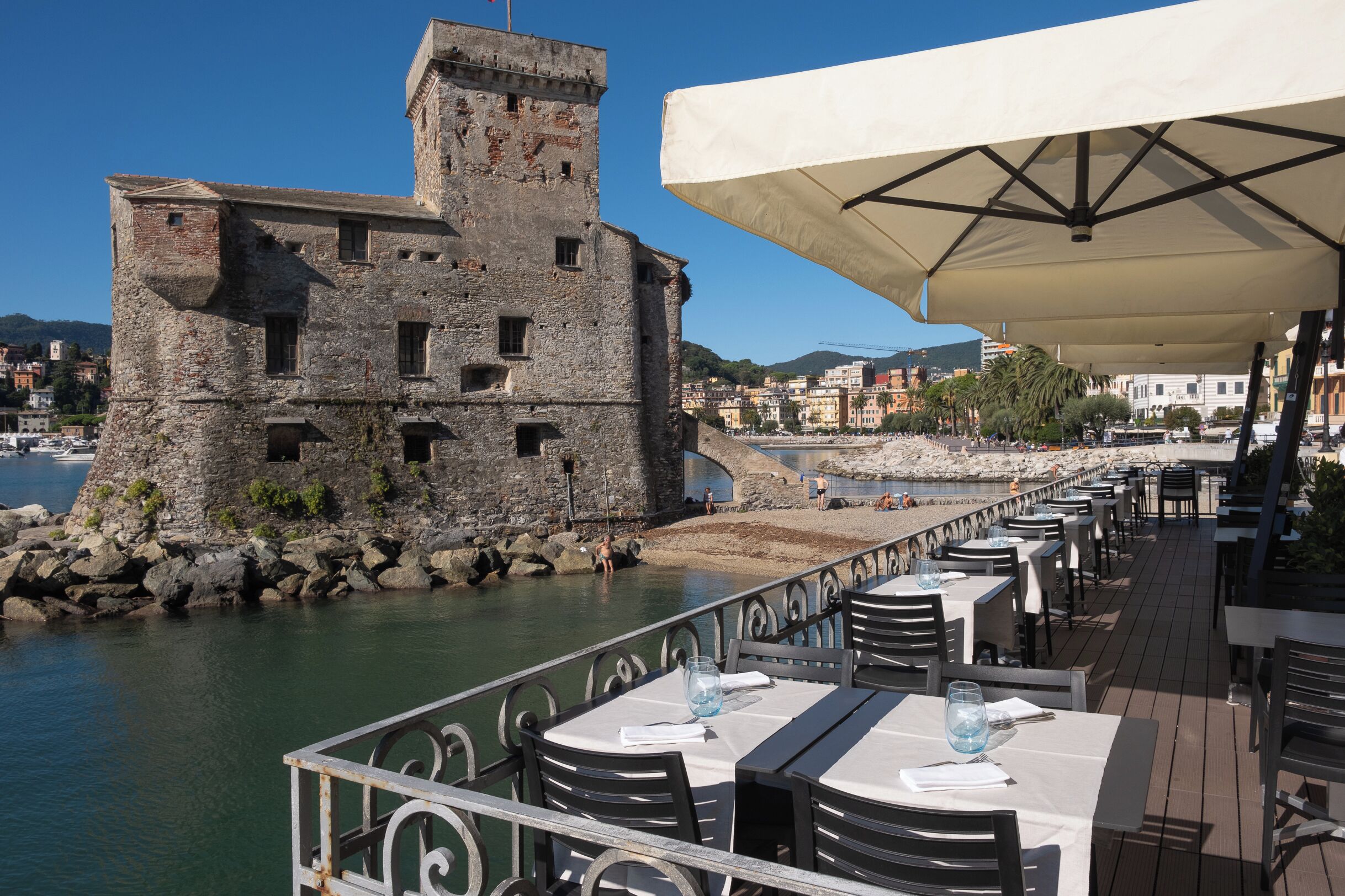 A seaside restaurant with outdoor seating overlooking a rocky waterfront and a historic building.