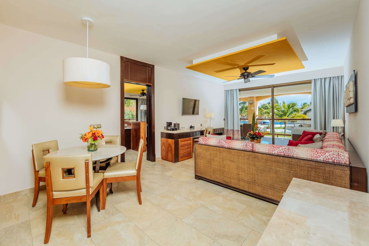 Habitación de hotel con comedor, sofá con vistas, ventilador de techo y ventana grande que da a una piscina.