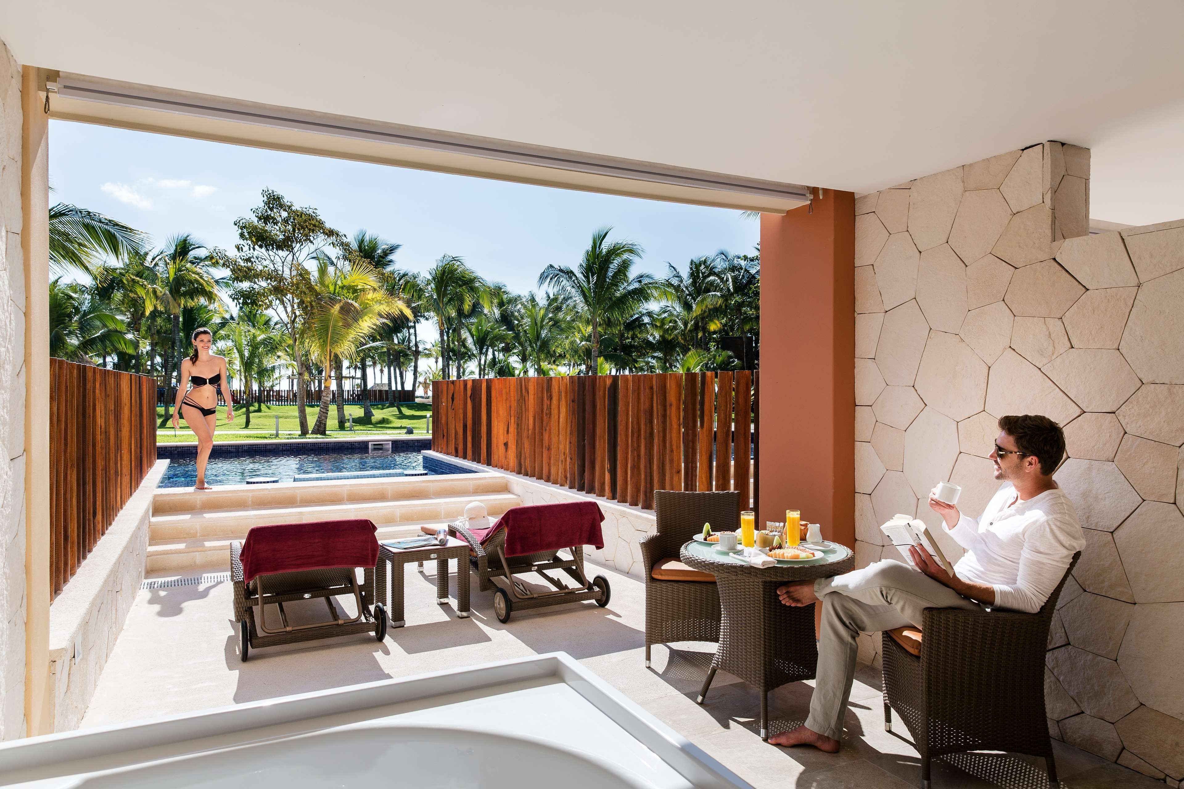 A poolside area with a lounge chair, a small table with a drink, and a person reading a book.
