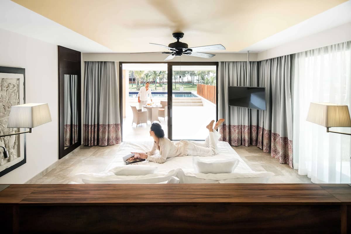 Habitación de hotel con cama, ventilador de techo, televisor y vistas a una piscina y zona de estar exterior.